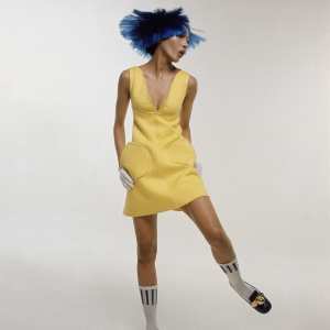 kellie-wilson-wearing-yellow-courrges-cotton-dress-plunged-at-the-neck-with-rounded-pockets-on-her-head-she-is-wearing-a-purple-dutch-boy-wig-and-on-her-feet-white-bobby-socks-and-blac1.jpg